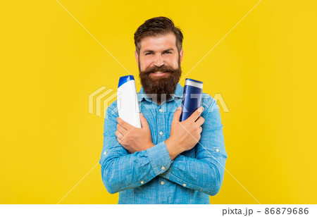 smiling brutal bearded man choosing hair conditioner bottle, presenting toiletries. 86879686