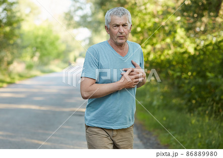 Photo of fatigue mature man feels pain in heart after jogging, has sullen expression, wears casual clothes, cares of his health. Handsome tired male has heart attack after active workout in open air 86889980