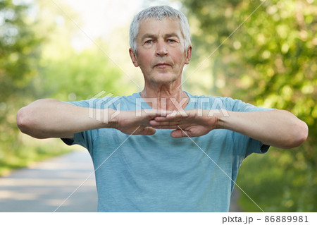 Confident grey haired male stretches hands, does physical exercise or workout in open air, has self assured facial expression stands outdoor, has healthy lifestyle. People, wellbeing and sport concept 86889981