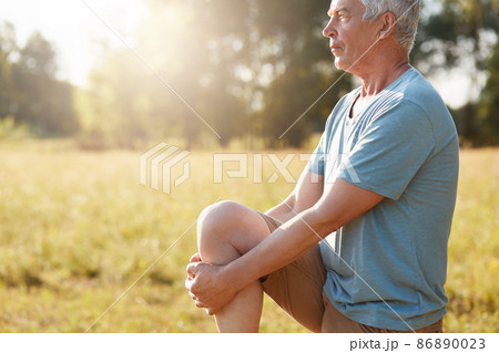 Cropped shot of serious mature male stretches legs on green field, looks into distance, does physical exercises in open air, feels fresh and healthy, dressed in casual t shirt, stands sideways Cropped shot of serious mature male stretches legs on green field, looks into distance, does physical exercises in open air, feels fresh and healthy, dressed in casual t shirt, stands sideways 86890023