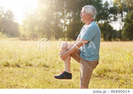 Sideways shot of healthy elderly male stretches legs, has serious expression, wears sport clothes and shoes, stands against nature background, does sport in open air. Physical activity concept Sideways shot of healthy elderly male stretches legs, has serious expression, wears sport clothes and shoes, stands against nature background, does sport in open air. Physical activity concept 86890024