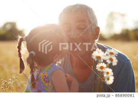 Close up shot of small grandchild embrace and kisses her grandfather who gives camomiles, have walk together in countryside, beautiful sunshine. Summer time concept. Pensioner with granddaughter 86890081