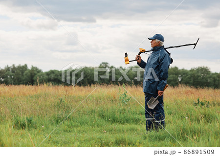 Side view of mature numismatist posing outdoors in field with shovel in hands and metal detector on his shoulder, looking in distance, treasure hunter in meadow. 86891509