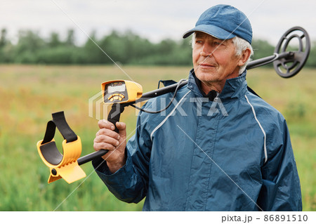 Senior numismatist wearing cap and jacket holding metal detector over his shoulder and looking away, treasure hunter searching historical artifacts. 86891510