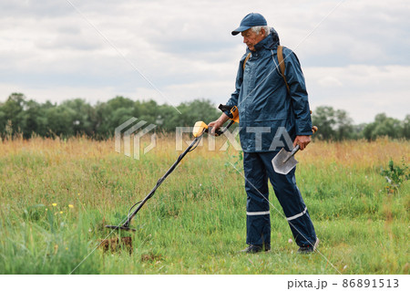 Full length outdoor portrait of numismatist in jacket and cap, holding in hands shovel and metal detector, looking for gold or historical artifacts in meadow. 86891513