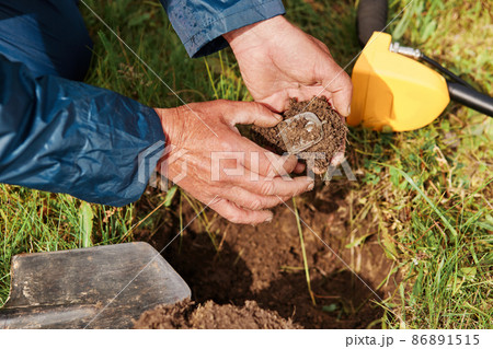 Unknown faceless treasure hunter holding his finding with ground in hands on background of grass, numismatist searching coins in field. 86891515