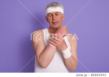 Horizontal shot of senior man dresses white sleeveless t shirt, hurts his wrist during sport training, white haired male posing isolated over lilac studio background. Fitness, health care concept. Horizontal shot of senior man dresses white sleeveless t shirt, hurts his wrist during sport training, white haired male posing isolated over lilac studio background. Fitness, health care concept. 86892644