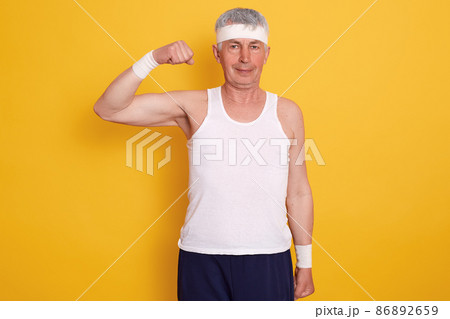 Indoor shot of senior man wearing sport clothing and headband, standing with one hand up and showing his biceps, being photographed after doing physical exercises. Healthy lifestyle concept. 86892659