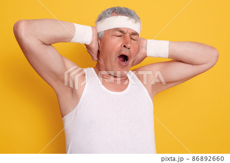 Indoor shot of yawning senior man keeping hands up, keeps mouth opened and eyes closed, standing against yellow wall, looks sleepy, wearing white t shirt and headband. Indoor shot of yawning senior man keeping hands up, keeps mouth opened and eyes closed, standing against yellow wall, looks sleepy, wearing white t shirt and headband. 86892660