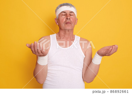 Closeup[ portrait of mature male with closed eyes, looks concentrated, posing with palms up, white haired man meditates at home while standing against yellow studio wall. Healthy lifestyle concept. Closeup[ portrait of mature male with closed eyes, looks concentrated, posing with palms up, white haired man meditates at home while standing against yellow studio wall. Healthy lifestyle concept. 86892661