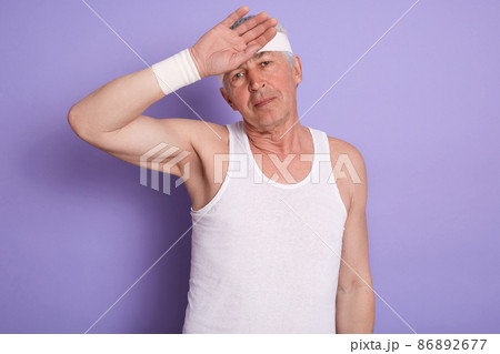 Portrait of exhausted determined senior man wiping out sweat from forehead with hand, having tired facial expression, doing sports, wearing headband and white shirt. People and lifestyle concept. Portrait of exhausted determined senior man wiping out sweat from forehead with hand, having tired facial expression, doing sports, wearing headband and white shirt. People and lifestyle concept. 86892677