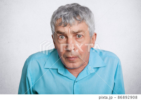 People, age and emotions concept. Surprised grey haired elderly man looks with unexpeted expression into camera, wears blue formal shirt, isolated over white background. Mature male poses indoor 86892908