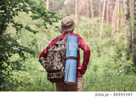 Back view of active backpacker looking for way out in forest, having sleeping pad and dark rucksack with thermos at his back, enjoying active holidays, stick to travel lifestyle. Travel concept. 86893485