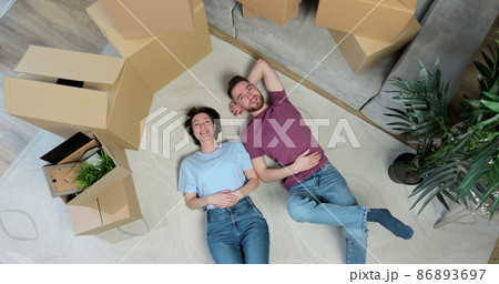 Happy but tired young couple lie on the floor of their new home. Young couple lying on floor of their new apartment in bedroom, talking and hugging. Carton boxes, carpet and bed are visible. Happy but tired young couple lie on the floor of their new home. Young couple lying on floor of their new apartment in bedroom, talking and hugging. Carton boxes, carpet and bed are visible. 86893697