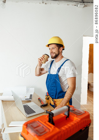 Relaxed handyman in uniform and hard hat drinking coffee during a break while doing renovation work indoors 86896082