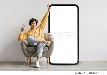Joyful guy sittiwng on chair near big empty smartphone screen, listening to music in wireless headphones over grey wall 86899655