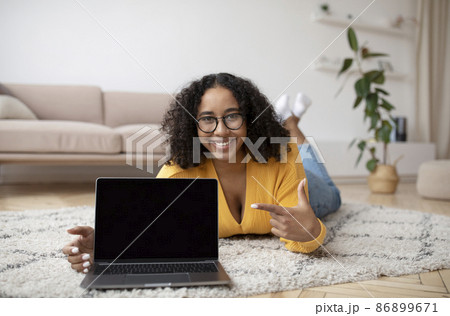 Cheery young black woman lying on floor, pointing at laptop computer with mockup for website design on screen 86899671