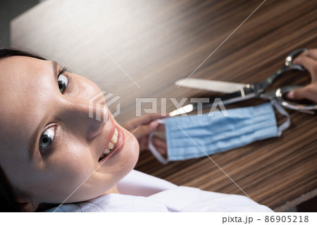 A young medical doctor is sitting at his desk and getting ready to cut the protective mask and thus end the ongoing pandemic and stringent restrictions on society. A young medical doctor is sitting at his desk and getting ready to cut the protective mask and thus end the ongoing pandemic and stringent restrictions on society. 86905218