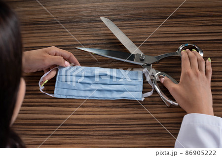 A young medical doctor is sitting at his desk and getting ready to cut the protective mask and thus end the ongoing pandemic and stringent restrictions on society. A young medical doctor is sitting at his desk and getting ready to cut the protective mask and thus end the ongoing pandemic and stringent restrictions on society. 86905222