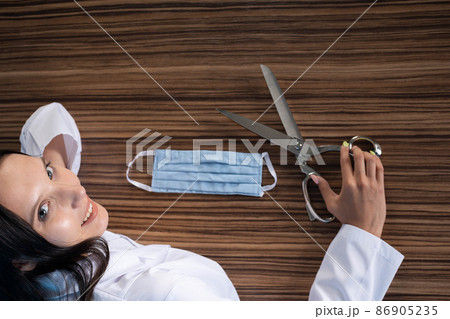 A young medical doctor is sitting at his desk and getting ready to cut the protective mask and thus end the ongoing pandemic and stringent restrictions on society. 86905235