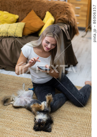 A dog is lying on the carpet and a girl is taking pictures of it with her smartphone. A multi-breed dog. 86906357