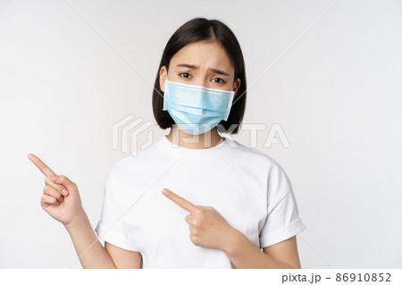Sad asian woman in medical mask, pointing fingers left, frowning and looking upset, complaining, demonstrating banner, standing over white background 86910852