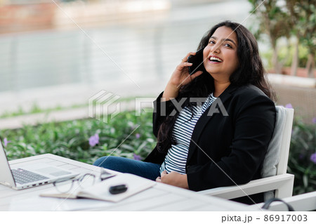 A young Indian woman is working on a laptop on the terrace in a cafe and talking on the phone. Online business, freelance. A young Indian woman is working on a laptop on the terrace in a cafe and talking on the phone. Online business, freelance. 86917003