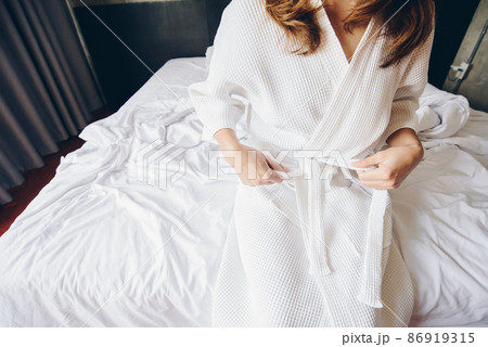 Cropped shot view of young woman sitting on the bed and tying up belt of white bathrobe before taking a bath or spa massage. 86919315