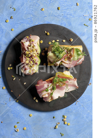 Three bruschettas with ham, micro-greenery and sprouts, on a black tarecla. Close-up on a blue background. Top view Three bruschettas with ham, micro-greenery and sprouts, on a black tarecla. Close-up on a blue background. Top view 86921762