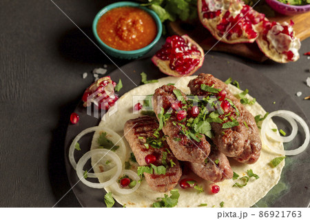 Sausages made of minced meat, on pita bread, with fresh herbs, onions, pomegranate seeds and hot tomato sauce. Close-up on a black background 86921763