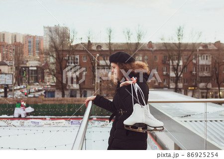 Young curly-haired woman in a beret with ice skates looks down at the ice rink, wants to learn how to skate Young curly-haired woman in a beret with ice skates looks down at the ice rink, wants to learn how to skate 86925254