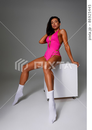 Beautiful sexy woman posing in pink fashionable swimsuit with smile on gray background in a studio on a cube, looking at camera Beautiful sexy woman posing in pink fashionable swimsuit with smile on gray background in a studio on a cube, looking at camera 86925445