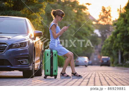 Young woman sitting on suitcase near her vehicle talking on her mobile phone on a city street in summer. Young woman sitting on suitcase near her vehicle talking on her mobile phone on a city street in summer. 86929881