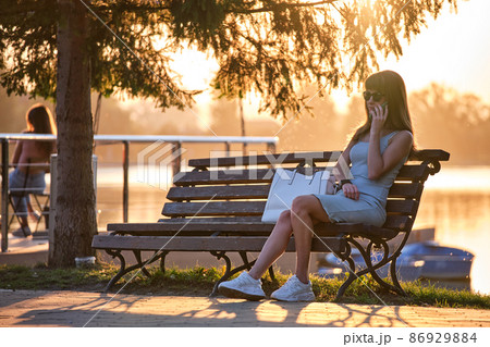Young woman sitting on park bench talking on her cellphone outdoors on warm summer evening. Communication and mobile connection concept. Young woman sitting on park bench talking on her cellphone outdoors on warm summer evening. Communication and mobile connection concept. 86929884