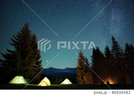 Silhouette of hikers resting besides burning bonfire near illuminated tourist tents on camping site in dark mountains under night sky with sparkling stars. Active lifestyle and outdoor living concept. Silhouette of hikers resting besides burning bonfire near illuminated tourist tents on camping site in dark mountains under night sky with sparkling stars. Active lifestyle and outdoor living concept. 86930482