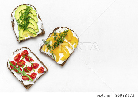 Variety of sandwiches for breakfast - slice of whole grain dark bread, yellow bell pepper, cream cheese, avocado, cherry tomatoes, garnished with dill, green arugula on white table Top view Flat lay Variety of sandwiches for breakfast - slice of whole grain dark bread, yellow bell pepper, cream cheese, avocado, cherry tomatoes, garnished with dill, green arugula on white table Top view Flat lay 86933973