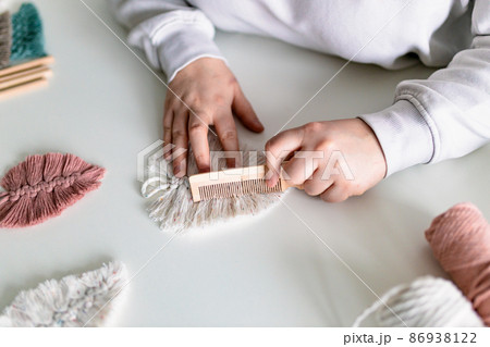 Woman working out last details of an almost-finished macrame piece, combing ropes inside a home room. Close up. Natural cotton threads and wooden beads. Female hobby. Wall hanging decor. Woman working out last details of an almost-finished macrame piece, combing ropes inside a home room. Close up. Natural cotton threads and wooden beads. Female hobby. Wall hanging decor. 86938122