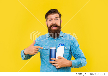 surprised brutal bearded man pointing finger on shampoo bottle on yellow background, skincare 86938904