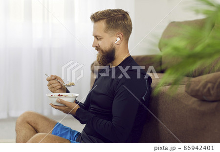 Athletic man after active morning sports training eats healthy food to restore energy. Side view of bearded man with wireless headphones in his ears sitting on floor near sofa with bowl of oatmeal. 86942401