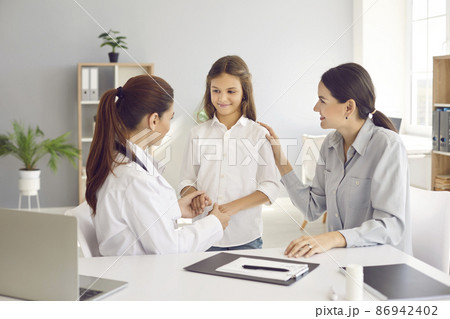 Little teen girl during medical examination receives support from her mother and friendly female pediatrician. Doctor holds child's hands and mother touches shoulder, assuring that everything is fine. 86942402