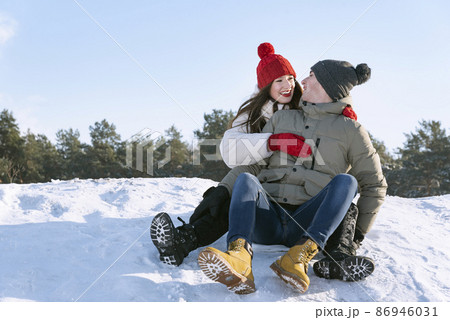 Young happy guy and a girl sit on the snow slide against the background of the forest and the blue sky Young happy guy and a girl sit on the snow slide against the background of the forest and the blue sky 86946031
