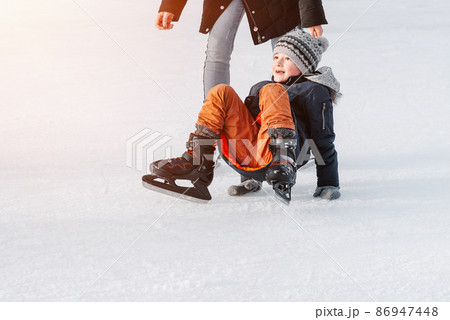 Soft,Selective focus.Mom with baby boy 6 years old, learn train, ride winter city rink, ice skating. The child gets up, fell on skates, kneels, play fun rest on weekend first steps child skates. 86947448