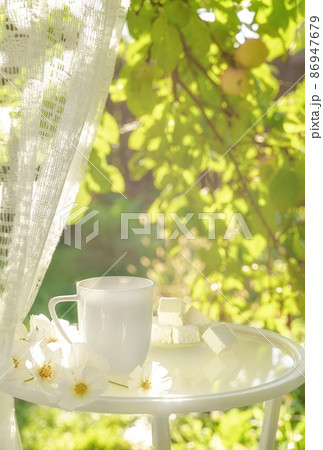 Beautiful porcelain coffee cup and Turkish delight sweets on glass table decorated with white flowers in summer garden in sunlight. Copy space. Summer drinks concept. 86947679