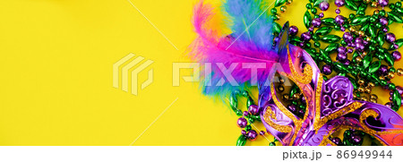 Carnival mask with feathers on yellow background. Multicolored beads in green, gold and purple. Mardi Gras or Fat Tuesday symbol. Copy space, banner format. Carnival mask with feathers on yellow background. Multicolored beads in green, gold and purple. Mardi Gras or Fat Tuesday symbol. Copy space, banner format. 86949944