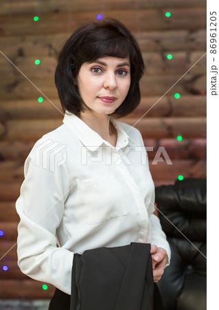 A brunette woman in a white blouse with a black jacket on her arm against a wooden wall with a garland 86961205