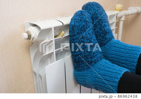 A woman warms her feet by the radiator on a cold winter day in blue woolen socks. Central heating system. Expensive heating costs during the cold season 86962438