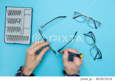 Master uses a screwdriver to tighten the screw in the frame of the glasses. Eyeglass frame repair. Optics salon and repair shop. Flat lay. Blue background. Master uses a screwdriver to tighten the screw in the frame of the glasses. Eyeglass frame repair. Optics salon and repair shop. Flat lay. Blue background. 86965816
