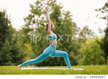 Slender focused girl performing yoga outside in early morning in park, standing in warrior pose 86969067