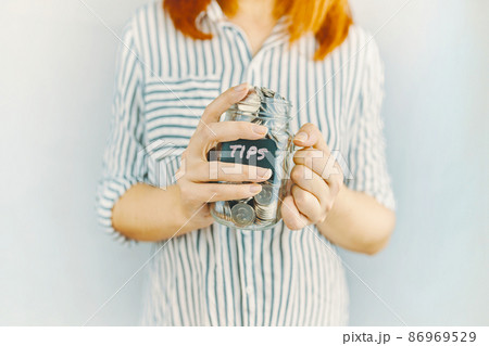 Glass bottle with silver coins in women's hands. Sticker with inscription on money jar. Redhead girl n white and black striped shirt holds her tips pot. Glass bottle with silver coins in women's hands. Sticker with inscription on money jar. Redhead girl n white and black striped shirt holds her tips pot. 86969529