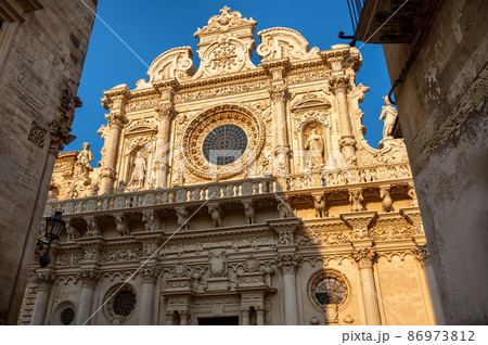 Lecce,Puglia,Italy.August 2021. The church of Santa Croce is the finest example of baroque. The facade illuminated by the warm light of the evening peeps out between two houses in the historic center Lecce,Puglia,Italy.August 2021. The church of Santa Croce is the finest example of baroque. The facade illuminated by the warm light of the evening peeps out between two houses in the historic center 86973812